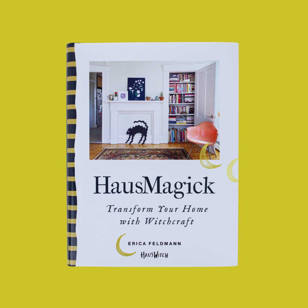 Cover of the book HausMagick: Transform Your Home with Witchcraft by Erica Feldmann, HausWitch. The photo above the title text shows a white-walled room with hardwood floors and an Oriental carpet. A white mantlepiece displays a decoration of an arched-back Halloween black cat. A closet full of colorful books and a peach-colored mid-century chair are to the right of the mantlepiece.