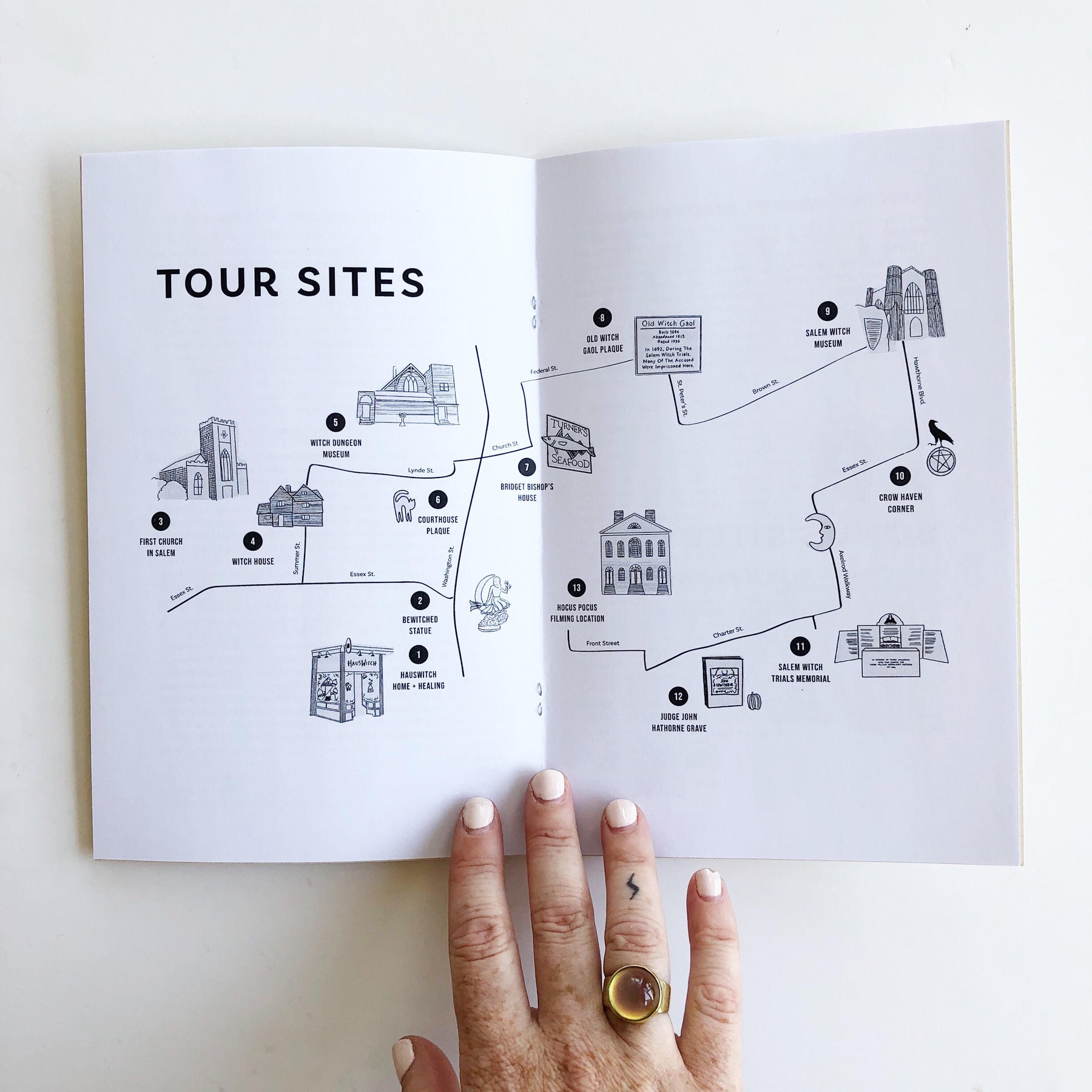 A hand with a lightning bolt tattoo and mood ring holds open a copy of Walking the Witch Trials: A DIY Tour of Salem History to show a map. Black line drawings of various tour sites in Salem include Salem Witch Museum, Hocus Pocus Filming Location, Bewitches Statue, and Salem Witch Trials Memorial.