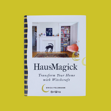 Load image into Gallery viewer, Cover of the book HausMagick: Transform Your Home with Witchcraft by Erica Feldmann, HausWitch. The photo above the title text shows a white-walled room with hardwood floors and an Oriental carpet. A white mantlepiece displays a decoration of an arched-back Halloween black cat. A closet full of colorful books and a peach-colored mid-century chair are to the right of the mantlepiece. 

