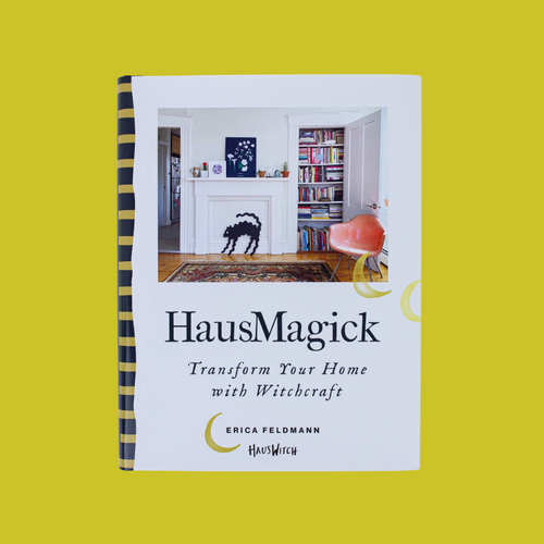 Cover of the book HausMagick: Transform Your Home with Witchcraft by Erica Feldmann, HausWitch. The photo above the title text shows a white-walled room with hardwood floors and an Oriental carpet. A white mantlepiece displays a decoration of an arched-back Halloween black cat. A closet full of colorful books and a peach-colored mid-century chair are to the right of the mantlepiece. 